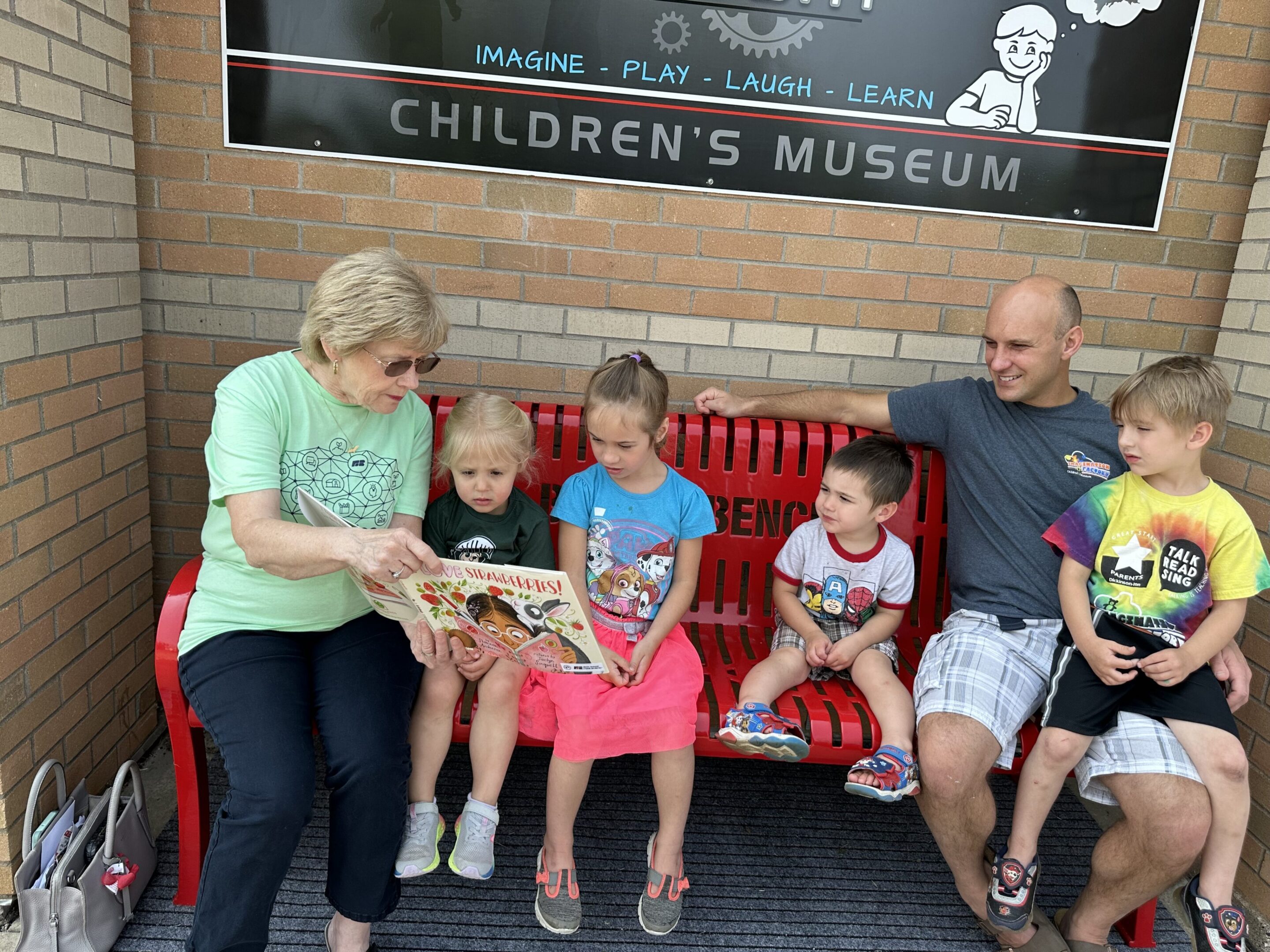 Elderly woman reading to children and an adult on a bench outside the Children's Museum.