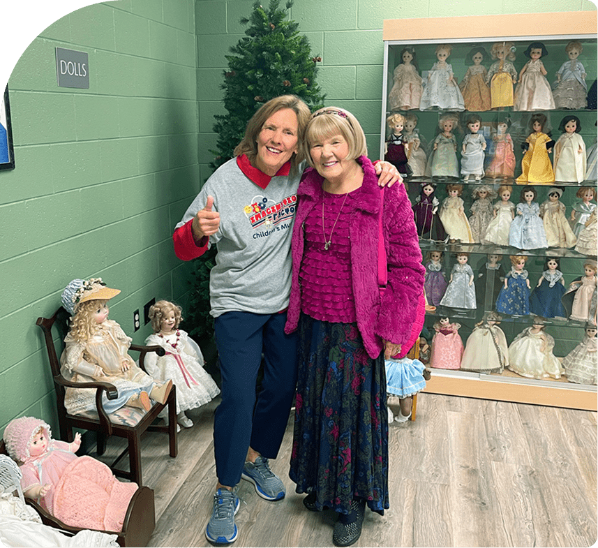 Two women stand in a room with a Christmas tree and a display case filled with vintage dolls.