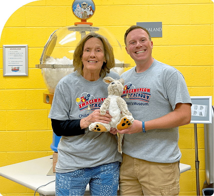 Two adults smiling and holding a stuffed kangaroo at a children’s museum.
