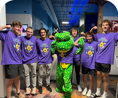 A group of young men in matching purple shirts pose with a green frog mascot indoors.