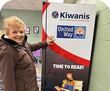 A smiling woman points to a Kiwanis and United Way reading banner.