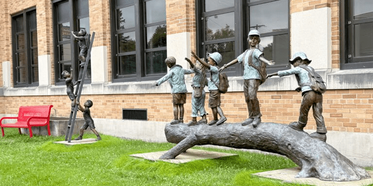 Bronze statue of children playing on a log outside a building.